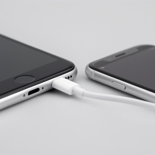 Câble Lightning blanc aligné près du port d’un smartphone compact de première génération, dans une mise en scène évoquant la compatibilité et le contrôle qualité.
