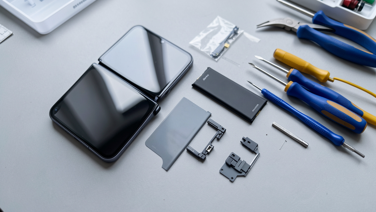 Smartphone pliable ouvert entouré d’un écran flexible, d’une batterie, d’une charnière de rechange et d’outils de réparation sur un établi propre.