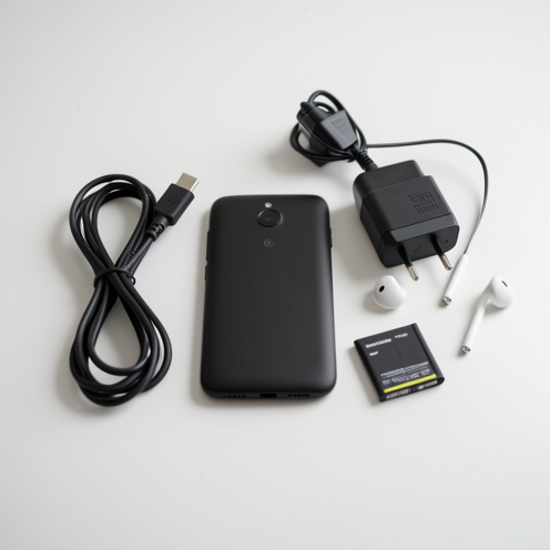 Smartphone compact noir de type Samsung Galaxy S5 Mini présenté avec son chargeur, ses écouteurs filaires et une batterie amovible sur une table blanche.
